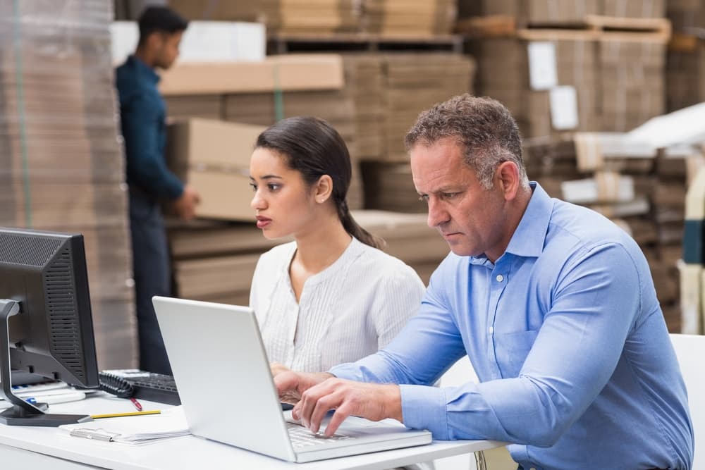 man on a laptop and women on a desktop computer, both looking at the screens, warehouse worker in the background building boxes