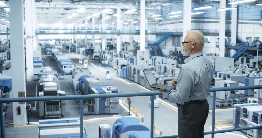 Senior manager with laptop overseeing automated manufacturing equipment in modern factory floor