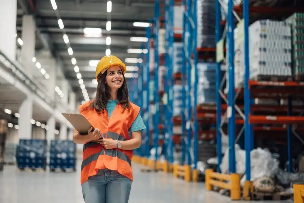 Female warehouse worker in safety vest and hard hat holding tablet in distribution center